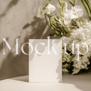 May include: A white card mockup with a shadow cast on a light beige surface. The card is positioned in front of a bouquet of white flowers, including orchids and hydrangeas.