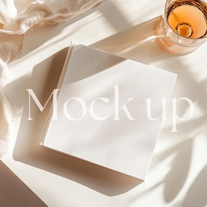 May include: A white book mockup on a light beige surface with a glass of amber liquid and a white fabric in the background. The text "Mock up" is written in white on the book.