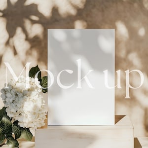 Puede incluir: Una maqueta de tarjeta blanca sobre una plataforma de madera con una flor de hortensia blanca en primer plano. El fondo es una pared marrón claro con sombras de la luz del sol.