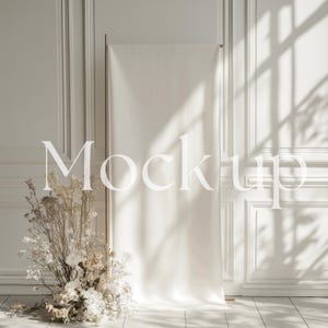 Può includere: Un banner di tessuto bianco con la parola "Mockup" in un carattere serif, esposto su un supporto dorato. Lo striscione è posto contro un muro bianco con modanature decorative. Una composizione floreale di fiori secchi e fiori bianchi è in primo piano.