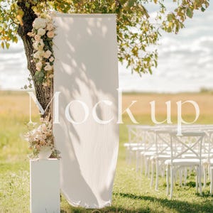 Può includere: Sfondo in tessuto bianco con un'arrangiamento floreale su un piedistallo bianco, allestito per una cerimonia di matrimonio all'aperto con sedie bianche sullo sfondo.