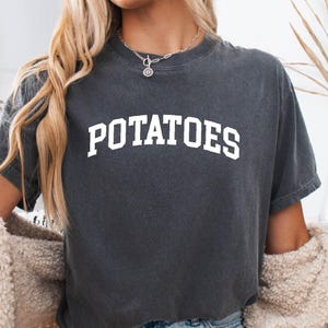 May include: Dark gray t-shirt with the word "POTATOES" in white, block letters. The shirt is worn with denim shorts and a cream-colored cardigan. A decorative pumpkin and dried plant are in the background.