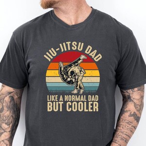 May include: A charcoal gray short-sleeve t-shirt featuring a graphic print of two figures performing a jiu-jitsu move. The design includes a sunset-colored circular background and the text JIU-JITSU DAD LIKE A NORMAL DAD BUT COOLER in beige.