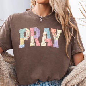 May include: A brown t-shirt with the word "PRAY" in colorful patchwork letters. The shirt is paired with denim shorts and a cream-colored cardigan. The image has a casual, relaxed vibe.