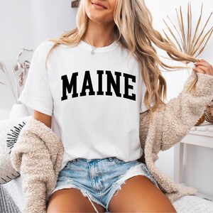 May include: A white short-sleeved t-shirt featuring the word MAINE printed in large, bold, black capital letters across the chest in an arched layout. The shirt has a crew neckline and is worn with light blue denim shorts and a beige cardigan.
