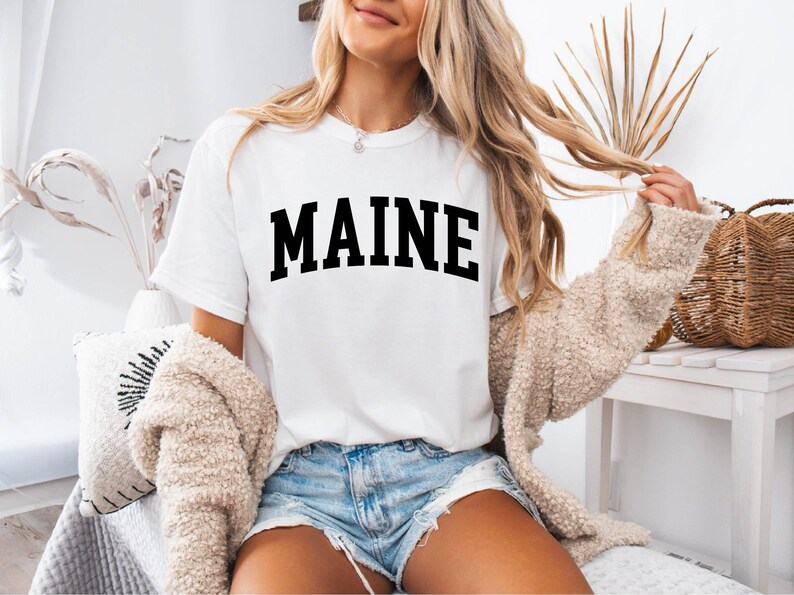 May include: A white short-sleeved t-shirt featuring the word MAINE printed in large, bold, black capital letters across the chest in an arched layout. The shirt has a crew neckline and is worn with light blue denim shorts and a beige cardigan.