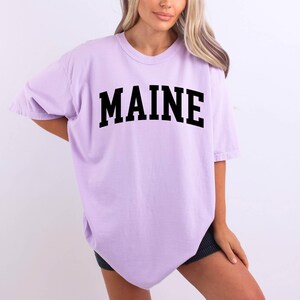 May include: A light purple short-sleeve t-shirt featuring the word MAINE printed in a bold, black, arched collegiate-style font across the chest. The garment has a crew neckline and a loose, relaxed fit with short sleeves and a straight hemline.