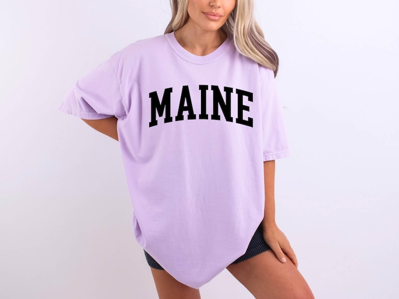 May include: A light purple short-sleeve t-shirt featuring the word MAINE printed in a bold, black, arched collegiate-style font across the chest. The garment has a crew neckline and a loose, relaxed fit with short sleeves and a straight hemline.