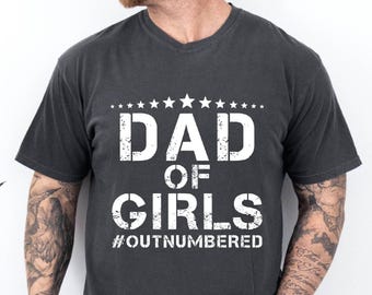 May include: Dark gray t-shirt with a V-neck featuring the text "DAD OF GIRLS" in distressed white lettering, with a row of white stars above the text. The hashtag "#OUTNUMBERED" is printed below the text.