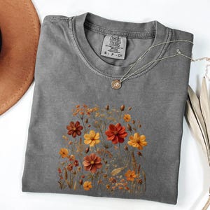 K&ouml;nnte beinhalten: Ein anthrazitfarbenes T-Shirt mit Rundhalsausschnitt und einem Grafikdruck verschiedener Wildblumen in Orange-, Gelb- und Rostt&ouml;nen. Das T-Shirt hat ein Comfort Colors-Markenetikett am Innenkragen mit der Gr&ouml;&szlig;enangabe S-CH sowie eine d&uuml;nne Goldkette.