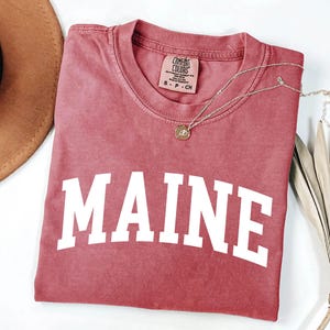 May include: A dusty rose short-sleeve t-shirt featuring the word MAINE printed in large white capital letters across the chest. The shirt has a crew neckline and a Comfort Colors brand label on the inside collar indicating size small.