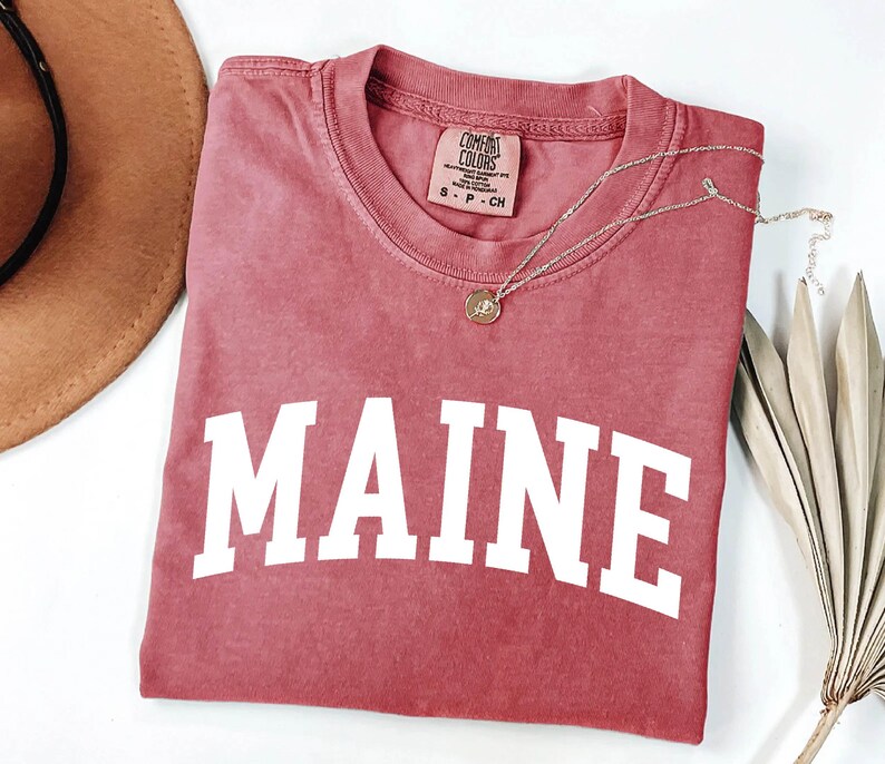 May include: A dusty rose short-sleeve t-shirt featuring the word MAINE printed in large white capital letters across the chest. The shirt has a crew neckline and a Comfort Colors brand label on the inside collar indicating size small.