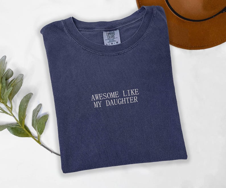May include: A navy blue t-shirt with the embroidered text "AWESOME LIKE MY DAUGHTER" in white. The shirt is folded and has a crew neck. A brown hat is partially visible in the background.