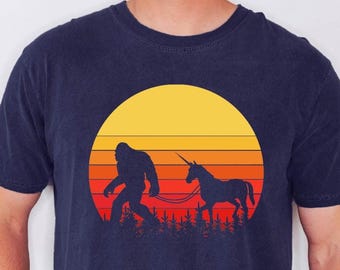May include: Navy blue t-shirt featuring a Bigfoot silhouette walking a unicorn on a leash. The design includes a sunset with yellow, orange, and red horizontal stripes. The graphic is set against a backdrop of trees.