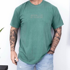 May include: A green t-shirt with the embroidered text "AWESOME LIKE MY DAUGHTERS". The shirt is a solid color and has a crew neck. The person wearing the shirt is also wearing blue jeans.