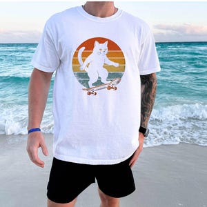 May include: White t-shirt with a retro-style graphic of a white cat skateboarding. The cat is wearing a red collar and is riding a skateboard with red wheels. The background is a sunset with orange, yellow, and red stripes.