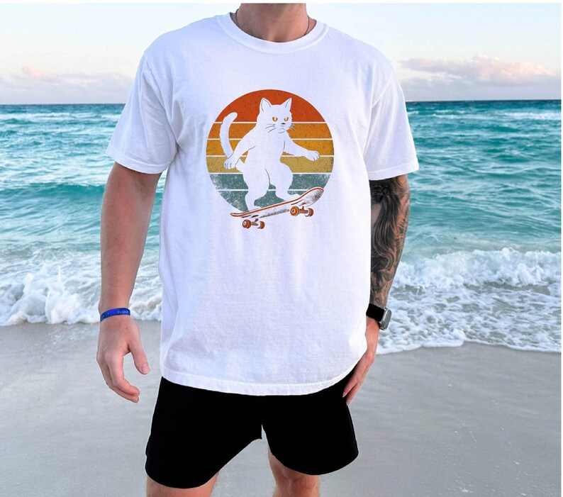 May include: White t-shirt with a retro-style graphic of a white cat skateboarding. The cat is wearing a red collar and is riding a skateboard with red wheels. The background is a sunset with orange, yellow, and red stripes.