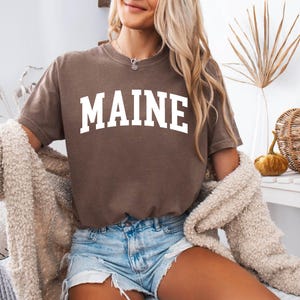 May include: A brown short-sleeved t-shirt featuring the word MAINE printed in large white capital letters across the chest in a classic collegiate-style arched font. The shirt is worn with light blue denim shorts and a beige textured cardigan.