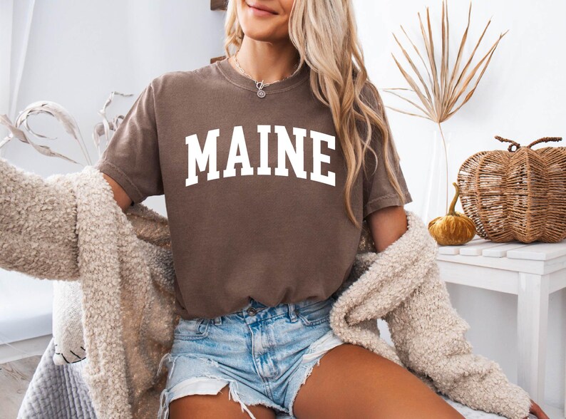 May include: A brown short-sleeved t-shirt featuring the word MAINE printed in large white capital letters across the chest in a classic collegiate-style arched font. The shirt is worn with light blue denim shorts and a beige textured cardigan.