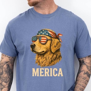 May include: A light blue t-shirt featuring a golden retriever wearing sunglasses and a bandana, both decorated with the American flag. The word "MERICA" is printed in a vintage font below the dog's image.