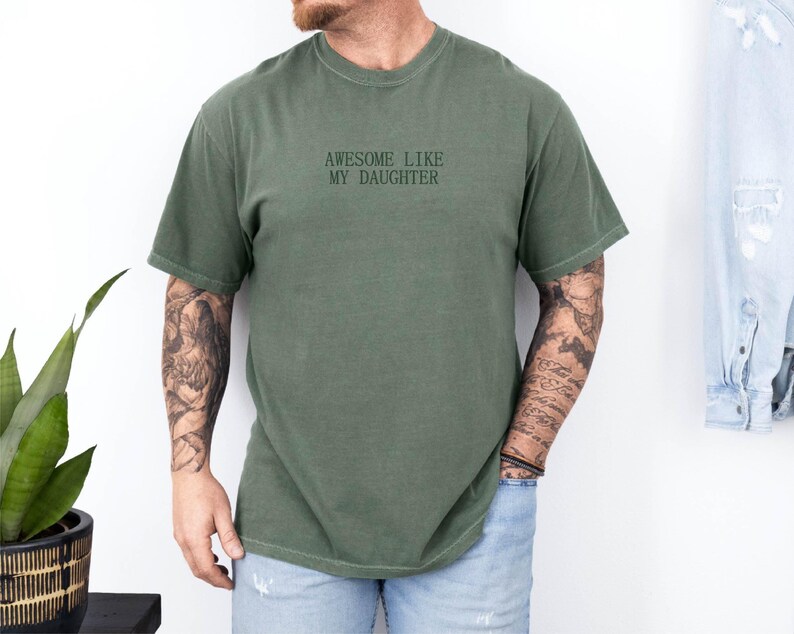 May include: Olive green t-shirt with the text "AWESOME LIKE MY DAUGHTER" in black. The shirt has short sleeves and a crew neck. The person wearing the shirt is also wearing blue jeans.