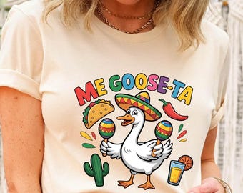 May include: Cream-colored t-shirt featuring a cartoon goose wearing a sombrero and holding maracas. The shirt has the text "ME GOOSE-TA" in colorful letters, along with images of a taco, chili pepper, cactus, and a drink.