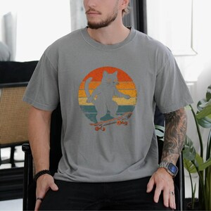 May include: A gray t-shirt with a vintage-style graphic of a cat skateboarding in front of a sunset. The cat is gray and the skateboard is brown.