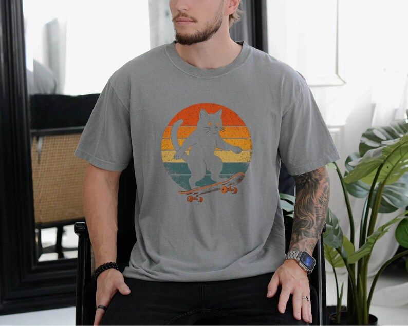 May include: A gray t-shirt with a vintage-style graphic of a cat skateboarding in front of a sunset. The cat is gray and the skateboard is brown.