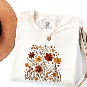 K&ouml;nnte beinhalten: Ein cremefarbenes Kurzarm-T-Shirt mit einem Comfort Colors-Etikett am Halsausschnitt und einem Grafikdruck verschiedener Wildblumen in Orange-, Gelb- und Braunt&ouml;nen. Eine d&uuml;nne goldfarbene Halskette mit einem kleinen runden Anh&auml;nger liegt auf dem T-Shirt.