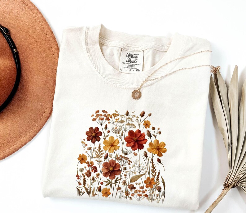 K&ouml;nnte beinhalten: Ein cremefarbenes Kurzarm-T-Shirt mit einem Comfort Colors-Etikett am Halsausschnitt und einem Grafikdruck verschiedener Wildblumen in Orange-, Gelb- und Braunt&ouml;nen. Eine d&uuml;nne goldfarbene Halskette mit einem kleinen runden Anh&auml;nger liegt auf dem T-Shirt.