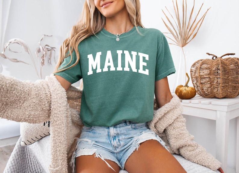 May include: A sage green short-sleeve t-shirt featuring the word MAINE printed in large white capital letters across the chest in a classic collegiate-style font. The garment has a crew neckline and a relaxed fit, paired with light blue denim shorts.