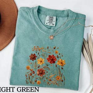 K&ouml;nnte beinhalten: Ein hellgr&uuml;nes Kurzarm-T-Shirt mit einem Grafikdruck verschiedener Wildblumen in Orange-, Rot- und Gelbt&ouml;nen. Das Shirt hat einen Rundhalsausschnitt und ein Comfort Colors Markenetikett am Innenkragen. Eine d&uuml;nne goldene Halskette liegt darauf.