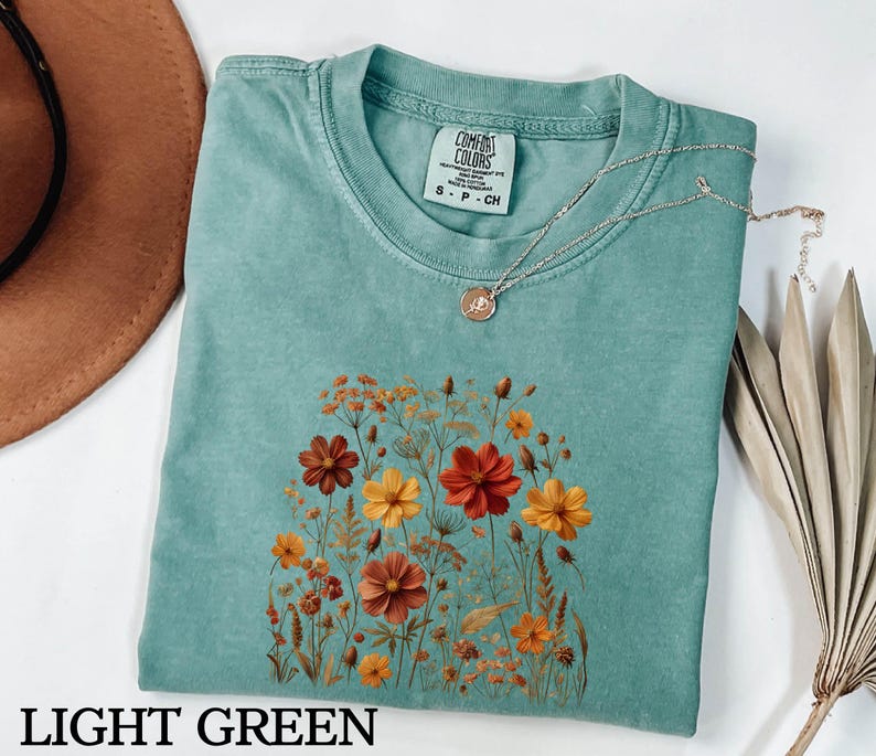 K&ouml;nnte beinhalten: Ein hellgr&uuml;nes Kurzarm-T-Shirt mit einem Grafikdruck verschiedener Wildblumen in Orange-, Rot- und Gelbt&ouml;nen. Das Shirt hat einen Rundhalsausschnitt und ein Comfort Colors Markenetikett am Innenkragen. Eine d&uuml;nne goldene Halskette liegt darauf.
