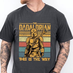 May include: Dark gray t-shirt featuring a graphic of a Mandalorian-style character with the text "THE DADALORIAN" and "THIS IS THE WAY" in a retro design. The character is in gold armor with a weapon.