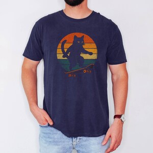 May include: A navy blue t-shirt with a retro sunset graphic featuring a black cat skateboarding. The cat has yellow eyes and is wearing a red collar.