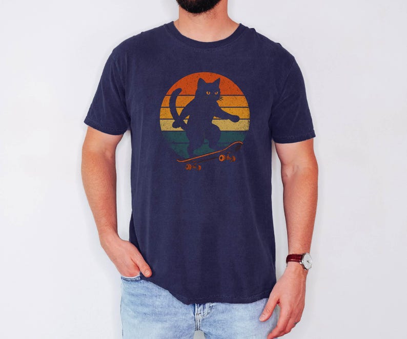 May include: A navy blue t-shirt with a retro sunset graphic featuring a black cat skateboarding. The cat has yellow eyes and is wearing a red collar.