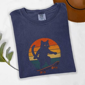 May include: A dark blue t-shirt with a retro-style graphic of a black cat skateboarding. The graphic features a sunset with orange, yellow, and red colors. The cat is wearing a skateboard with the word "O.S.Y" printed on it.