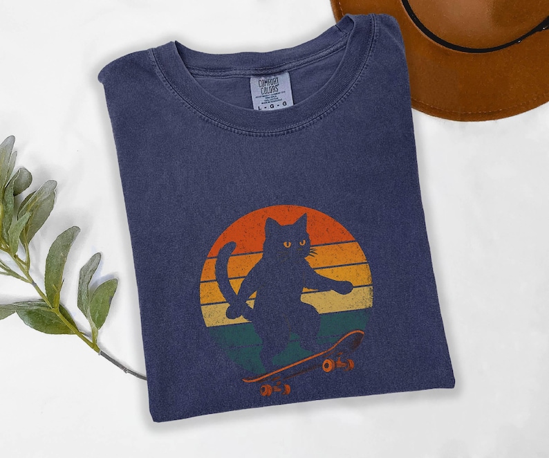 May include: A dark blue t-shirt with a retro-style graphic of a black cat skateboarding. The graphic features a sunset with orange, yellow, and red colors. The cat is wearing a skateboard with the word "O.S.Y" printed on it.