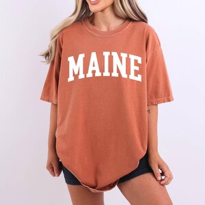 May include: A rust-colored, short-sleeved t-shirt featuring the word MAINE printed in large, white, block-style capital letters across the chest. The garment has a crew neckline and a relaxed, oversized fit with a slightly faded, vintage-inspired texture.
