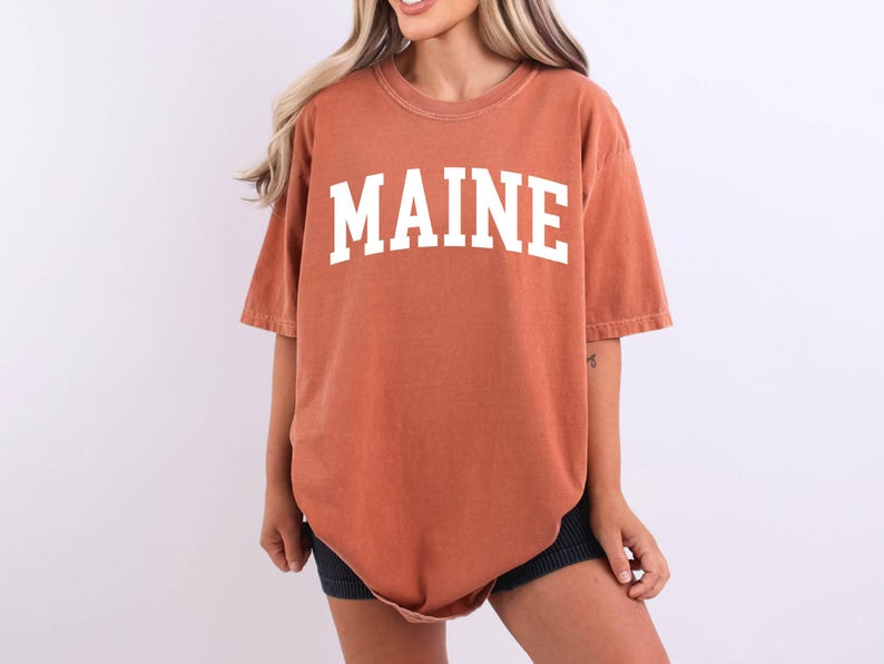 May include: A rust-colored, short-sleeved t-shirt featuring the word MAINE printed in large, white, block-style capital letters across the chest. The garment has a crew neckline and a relaxed, oversized fit with a slightly faded, vintage-inspired texture.