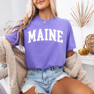 May include: A purple short-sleeved t-shirt featuring the word MAINE printed in large white capital letters across the chest in an arched font. The shirt has a crew neckline and a relaxed fit, paired with light blue denim shorts with frayed edges.