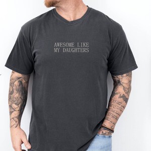 May include: Dark gray t-shirt with the text "AWESOME LIKE MY DAUGHTERS" in a simple, sans-serif font. The shirt has a crew neck and short sleeves. The model is wearing blue jeans.