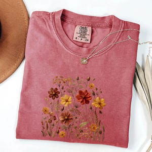 K&ouml;nnte beinhalten: Ein altrosa T-Shirt mit kurzen &Auml;rmeln und einem Grafikdruck verschiedener Wildblumen in Orange-, Gelb- und Braunt&ouml;nen. Das T-Shirt hat einen Rundhalsausschnitt und ein Comfort Colors Markenetikett am Innenkragen mit der Gr&ouml;&szlig;enangabe S-CH.