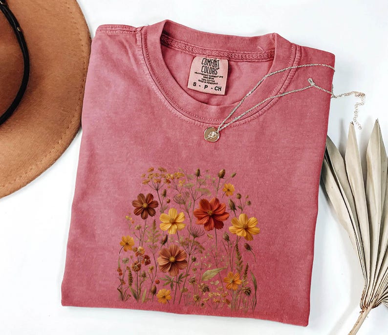 K&ouml;nnte beinhalten: Ein altrosa T-Shirt mit kurzen &Auml;rmeln und einem Grafikdruck verschiedener Wildblumen in Orange-, Gelb- und Braunt&ouml;nen. Das T-Shirt hat einen Rundhalsausschnitt und ein Comfort Colors Markenetikett am Innenkragen mit der Gr&ouml;&szlig;enangabe S-CH.