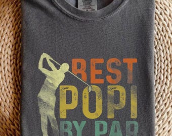 May include: Dark gray t-shirt with the words "BEST POPI BY PAR" in orange, yellow, and green. A golfer is depicted in a yellow and green silhouette. The shirt is folded on a woven surface.