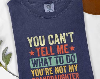 May include: A navy blue t-shirt with the text "YOU CAN'T TELL ME WHAT TO DO YOU'RE NOT MY GRANDDAUGHTER" in yellow, red, and green. The shirt has a crew neck and a Comfort Colors tag.