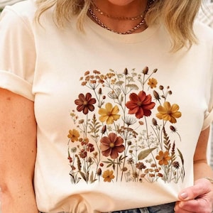 K&ouml;nnte beinhalten: Ein cremefarbenes Kurzarm-T-Shirt mit einem zentralen Grafikdruck verschiedener Wildblumen in Rost-, Orange-, Gelb- und Braunt&ouml;nen. Die botanische Illustration zeigt detaillierte St&auml;ngel, Bl&auml;tter und bl&uuml;hende Blumenk&ouml;pfe.