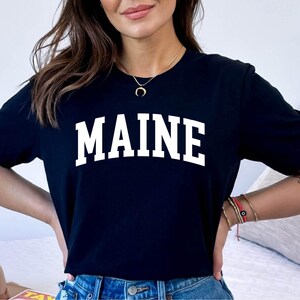 May include: A black short-sleeved t-shirt featuring the word MAINE printed in large, white, arched capital letters across the chest. The garment has a classic crew neckline and a straight hem, providing a simple and clean look for casual wear.