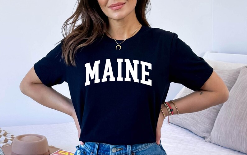May include: A black short-sleeved t-shirt featuring the word MAINE printed in large, white, arched capital letters across the chest. The garment has a classic crew neckline and a straight hem, providing a simple and clean look for casual wear.