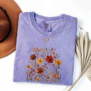 K&ouml;nnte beinhalten: Ein lavendelfarbenes Kurzarm-T-Shirt mit einem Comfort Colors-Etikett am Halsausschnitt. Das Shirt zeigt einen Grafikdruck verschiedener Wildblumen in Orange-, Gelb- und Braunt&ouml;nen. Eine goldfarbene Halskette mit einem kleinen runden Anh&auml;nger liegt auf dem Stoff.